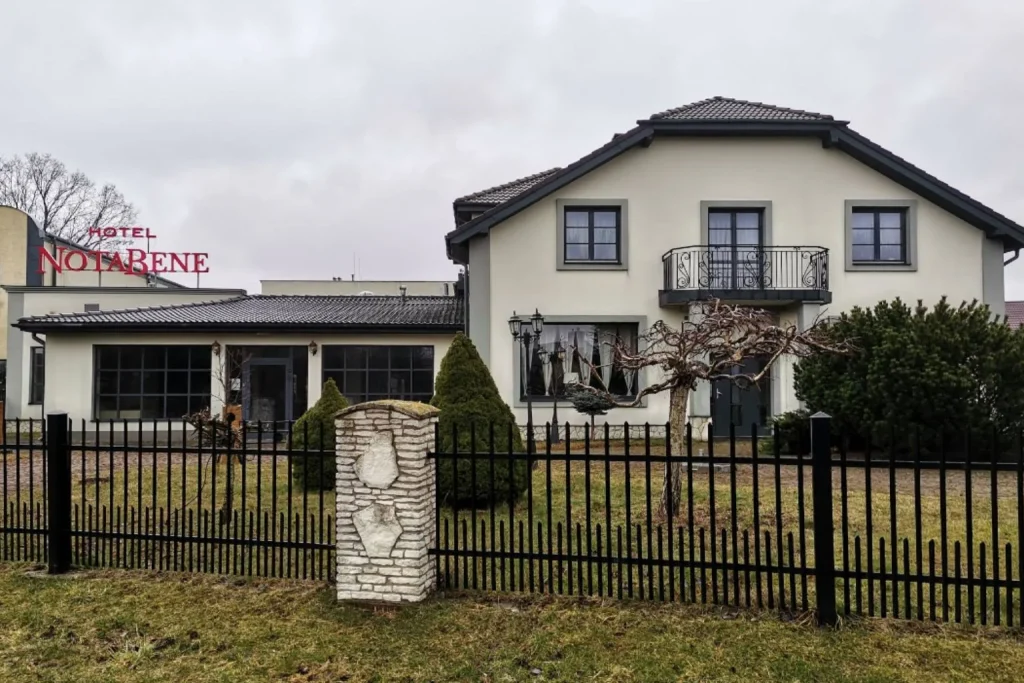 Front exterior of Hotel Notabene in Świdnik, Poland, featuring a modern cream-colored building with landscaped gardens, a black wrought-iron fence, and the hotel entrance.